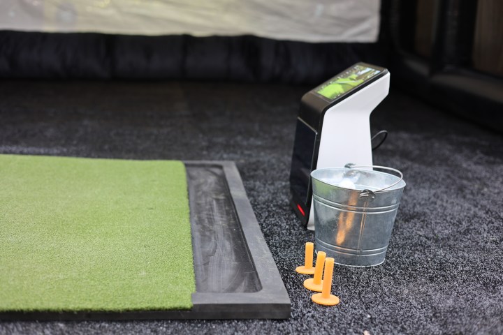 Golf mat with a metal bucket, tees, and an electronic device on a carpeted floor.