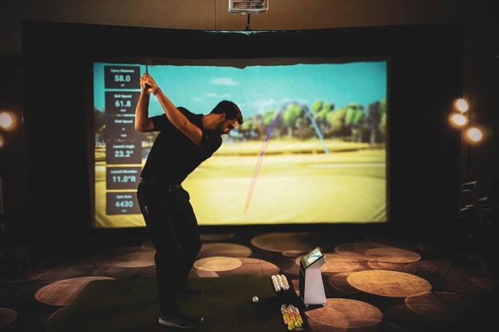 Person swings golf club in indoor simulator with data on screen.
