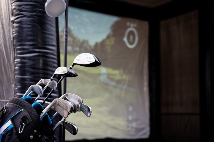 Golf clubs in a bag with an indoor golf simulator screen displaying a course in the background.