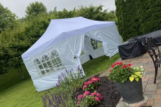 White tent on lawn near flower beds and patio with trees in background.